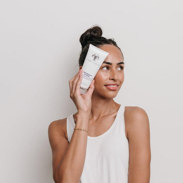 A woman in a white tank top holding a tube of Tidal Moisture for Body | Circadia skincare product near her face, smiling softly against a plain background.