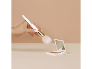 A hand holding the Bronzing Powder Brush | NUDA with a white handle and gold ferrule, poised above an open compact powder on a beige surface against a neutral background.