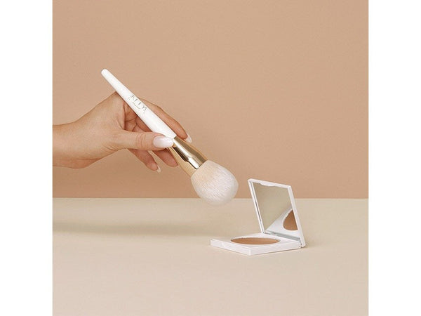 A hand holding the Bronzing Powder Brush | NUDA with a white handle and gold ferrule, poised above an open compact powder on a beige surface against a neutral background.