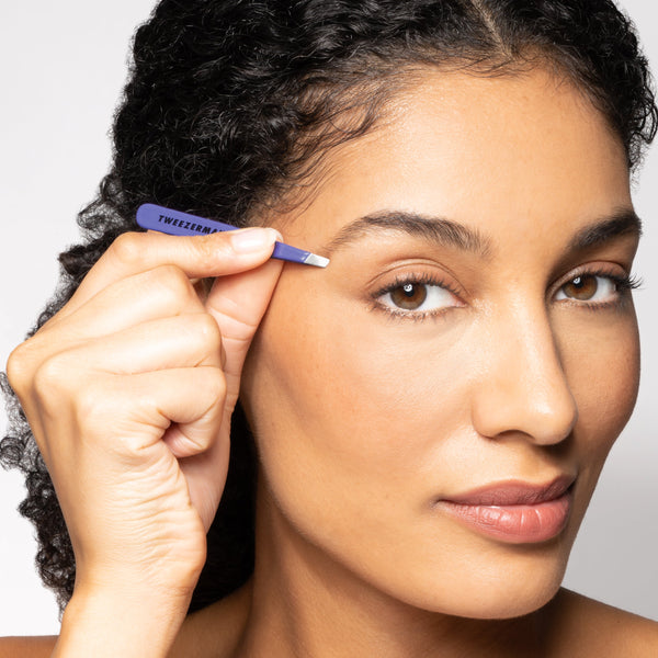 Woman using Blooming Lilac Mini Slant Tweezer | Tweezerman to groom her eyebrow, demonstrating precise skincare and beauty care application.
