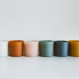 A row of minimalist colored glass jar candles including white, amber, pale pink, sage green, slate blue, and mustard yellow, featuring Vetiver + Tonka Dignity Series Glass Jar Soy Candle | Calyan Wax Co. - 5.3 oz.