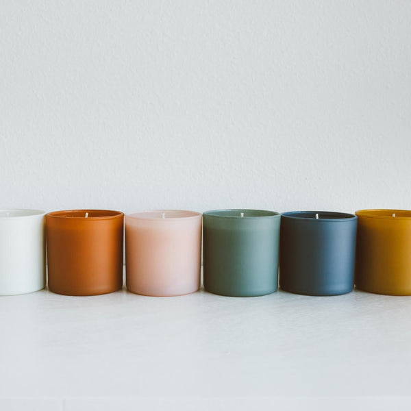 A row of minimalist colored glass jar candles including white, amber, pale pink, sage green, slate blue, and mustard yellow, featuring Vetiver + Tonka Dignity Series Glass Jar Soy Candle | Calyan Wax Co. - 5.3 oz.