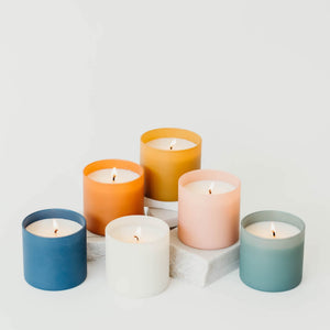 A collection of six lit candles in pastel-colored glass jars arranged on white blocks, featuring the Vetiver + Tonka Dignity Series Glass Jar Soy Candle | Calyan Wax Co. - 5.3 oz.