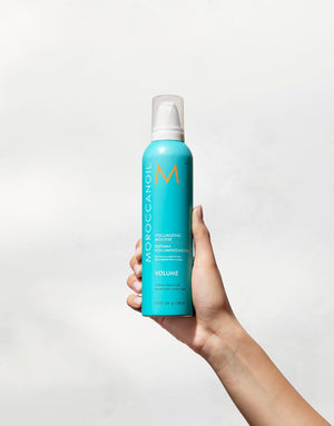 Hand holding a blue bottle of Volumizing Mousse | Moroccanoil with a white pump cap against a plain white background.