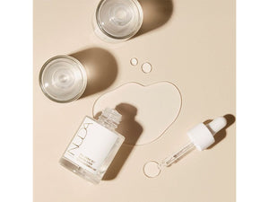 Clear glass bottle of Hyaluronic Self Tanning Serum | NUDA with a white dropper cap, spilled serum droplets, and two clear glass cups on a beige background, showcasing skincare and body care wellness.