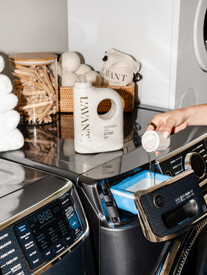 A person pouring High Performing Laundry Detergent - Fresh Linen | L'AVANT Collective into a washing machine detergent drawer in a modern laundry room with neatly stacked towels and natural laundry accessories.