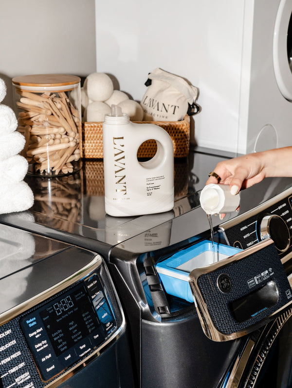 A person pouring High Performing Laundry Detergent - Fresh Linen | L'AVANT Collective into a washing machine detergent drawer in a modern laundry room with neatly stacked towels and natural laundry accessories.