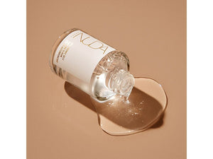 Clear glass bottle of Hyaluronic Self Tanning Serum | NUDA spilling a light tan liquid on a beige surface, highlighting skincare and body care wellness.