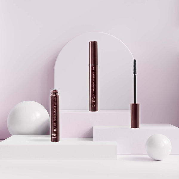 Three sleek brown tubes of Lash Extension Tubing Mascara | Blinc displayed on white geometric stands with a soft pink background and white spherical decor.