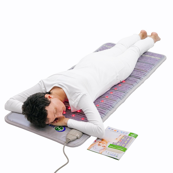 Person lying face down using the Platinum-Mat™ Full Short 6024 Firm - Photon Advanced PEMF InfraMat Pro® | HealthyLine emitting red light for wellness and recovery on a gray mat with white clothing and an instruction booklet nearby.