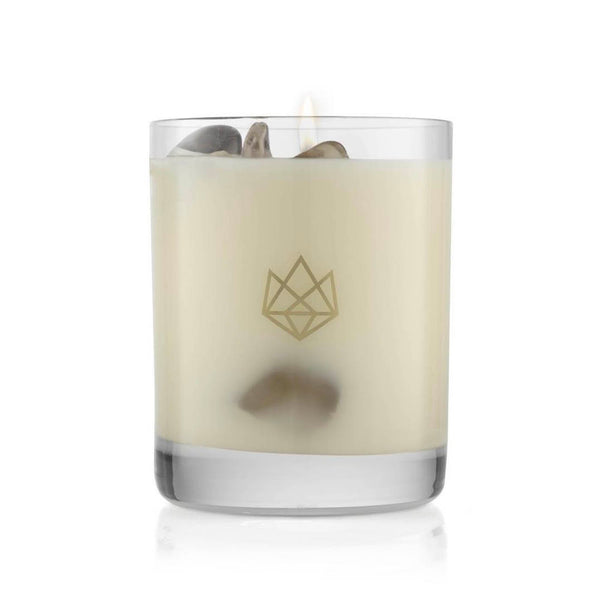 Lit creamy white candle with small decorative stones inside a clear glass holder featuring a subtle geometric logo, Defense Glass Candle | Aluminate Life.
