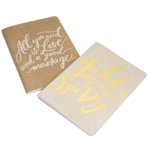 Two small notebooks with hand-lettered covers, one reading "All you need is love and a good massage notebook | Scout Books," the other with yellow script saying "You had me at Spa Day.