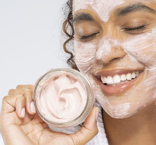 Smiling woman with a creamy pink facial mask applied, holding an open jar of Mighty Brighty™ Vitamin C + Chamomile Brightening Mask | Farmhouse Fresh, showcasing skincare and wellness.