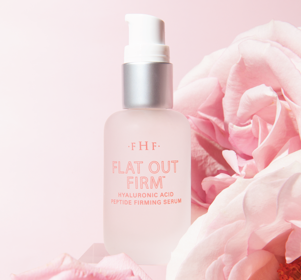 Bottle of Flat Out Firm™ Hyaluronic Acid Peptide Firming Serum | Farmhouse Fresh on a pastel pink background surrounded by soft pink flowers, highlighting skincare and beauty.