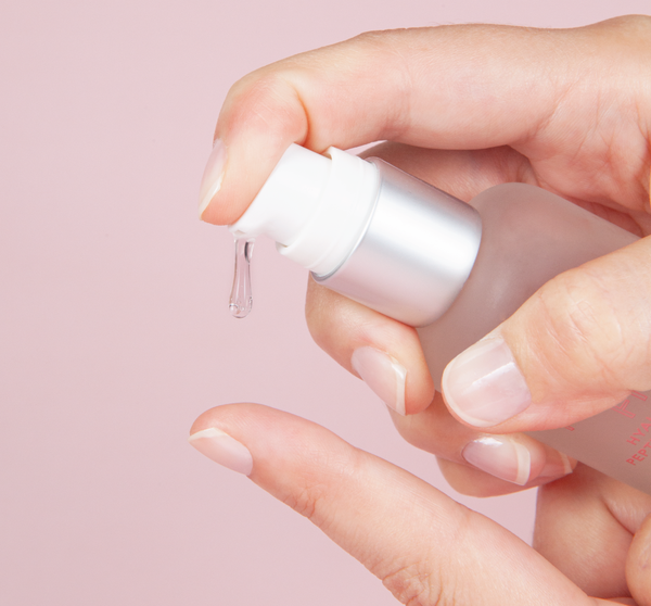Hand dispensing a clear serum drop from a frosted bottle with silver pump against a soft pink background for Flat Out Firm™ Hyaluronic Acid Peptide Firming Serum | Farmhouse Fresh skincare.
