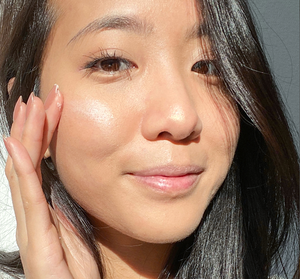 Young woman with radiant, glowing skin gently touching her cheek, showcasing the effects of Full Moon Dip® Iridescent Illumination Ageless Facial Mousse with Peptides + Retinol | Farmhouse Fresh skincare product.