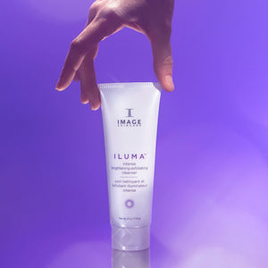 Hand holding a tube of ILUMA intense brightening exfoliating cleanser | IMAGE Skincare against a soft purple background, highlighting skincare and wellness products.