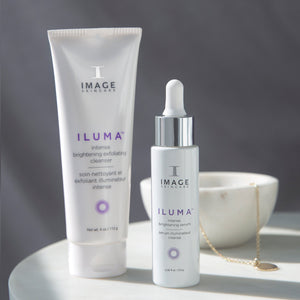 A white tube and a white dropper bottle of ILUMA® intense brightening serum | IMAGE Skincare displayed on a white surface with soft natural lighting and a blurred white bowl with a gold necklace in the background.