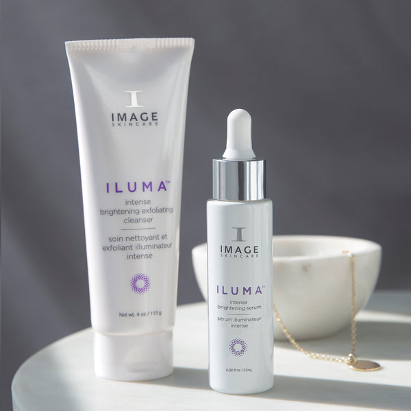 A white tube and a white dropper bottle of ILUMA® intense brightening serum | IMAGE Skincare displayed on a white surface with soft natural lighting and a blurred white bowl with a gold necklace in the background.