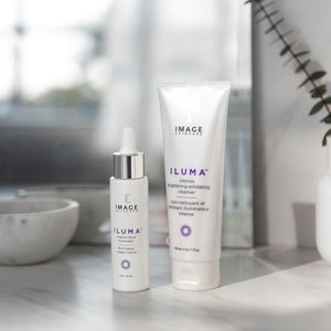 Two IMAGE Skincare products from the ILUMA line, including an intense facial illuminator dropper bottle and brightening exfoliating cleanser tube, displayed on a marble surface with soft natural light, promoting skincare and wellness.