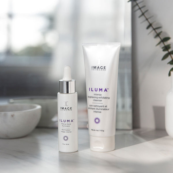 Two IMAGE Skincare products from the ILUMA line, including an intense facial illuminator dropper bottle and brightening exfoliating cleanser tube, displayed on a marble surface with soft natural light, promoting skincare and wellness.
