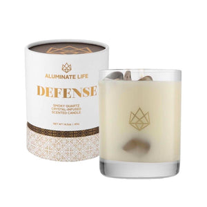Defense Glass Candle | Aluminate Life featuring a creamy white candle in a clear glass holder with smoky quartz crystals inside, next to its white and gold decorative packaging.