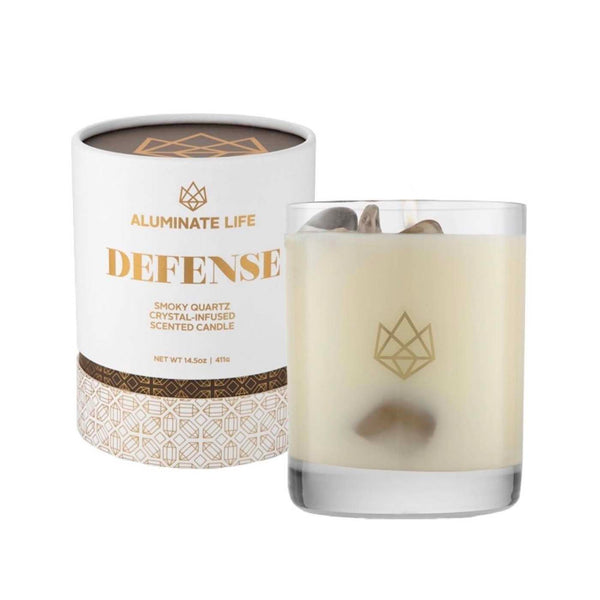 Defense Glass Candle | Aluminate Life featuring a creamy white candle in a clear glass holder with smoky quartz crystals inside, next to its white and gold decorative packaging.