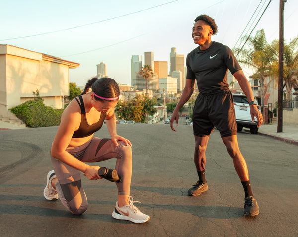 Woman using the Hypervolt 2 Pro | Hyperice massage device on her thigh outdoors while a man stands nearby smiling during a fitness session in the city.