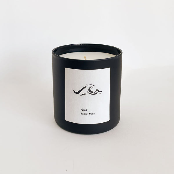 Minimalist black candle container with a white label featuring a wave design and text, showcasing No. 4 Sunset Swim (Amalfi Coast) Candle | Lucky Owl for a calming wellness ambiance.