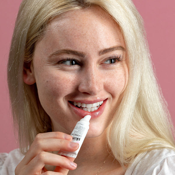A smiling woman with blonde hair applies ORMEDIC sheer pink lip enhancement complex | IMAGE Skincare against a soft pink background, highlighting skincare and wellness.