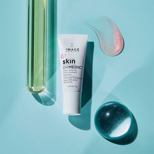 Tube of ORMEDIC sheer pink lip enhancement complex | IMAGE Skincare on a blue background with a clear pink gel swatch, a green liquid-filled glass tube, and a reflective round glass orb.