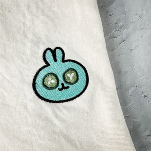 Close-up of a soft ivory fabric featuring a small embroidered turquoise bunny with green eyes on the Spa Bunny Comfy Tee | Lucky Owl, emphasizing cozy wellness style.