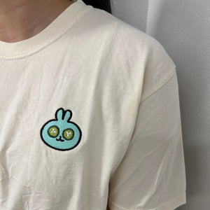 Close-up of a person wearing an ivory Spa Bunny Comfy Tee | Lucky Owl featuring a small, embroidered blue bunny with green eyes on the chest, made from soft comfortable fabric.