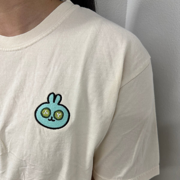 Close-up of a person wearing an ivory Spa Bunny Comfy Tee | Lucky Owl featuring a small, embroidered blue bunny with green eyes on the chest, made from soft comfortable fabric.