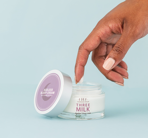 A hand dipping a finger into a jar of Three Milk Ageless Sleep Cream with Peptides | Farmhouse Fresh against a light blue background.