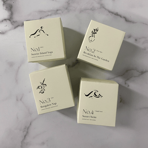 Four cream-colored candle boxes arranged on a marble surface, including one labeled No. 3 Bungalow Nap (Fiji) Candle | Lucky Owl with minimalist black botanical and yoga illustrations.