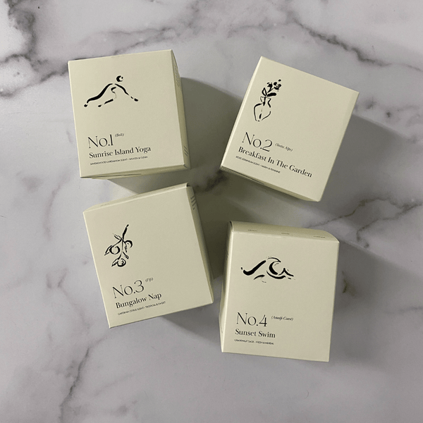 Four cream-colored candle boxes arranged on a marble surface, including one labeled No. 3 Bungalow Nap (Fiji) Candle | Lucky Owl with minimalist black botanical and yoga illustrations.