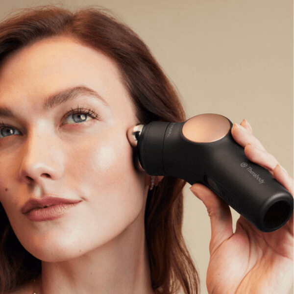A woman using the Theraface PRO | Therabody wellness device on her cheek, demonstrating a skincare and recovery tool for facial care and relaxation.