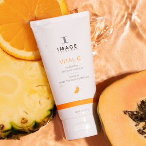 VITAL C hydrating enzyme masque | IMAGE Skincare tube surrounded by fresh pineapple, papaya, and orange slices on a shiny bronze surface, highlighting a tropical skincare theme.