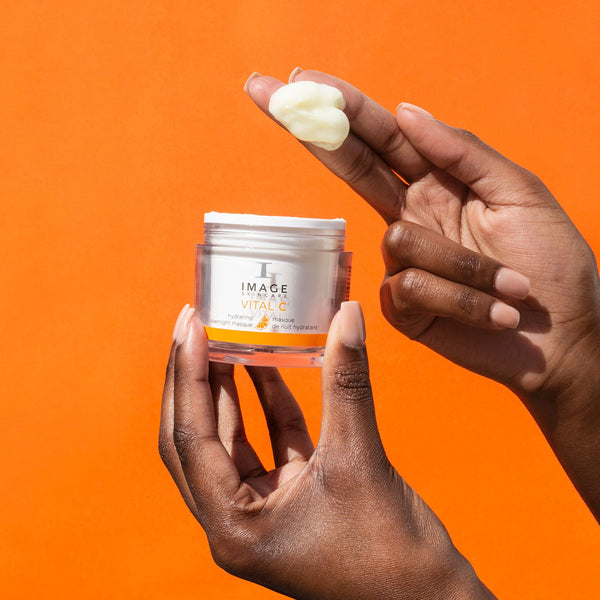 Hands holding an open jar and a dollop of VITAL C hydrating overnight masque | IMAGE Skincare against a vibrant orange background, showcasing the creamy texture of the skincare product.