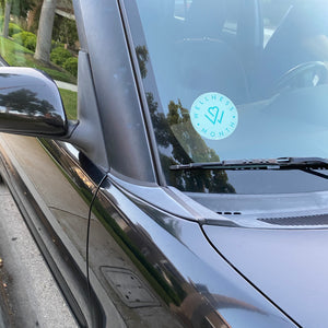 Car windshield with a circular blue and white Wellness Month Window Cling | Wellness Month decal placed on the inside near the windshield wiper.