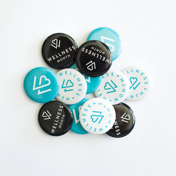 A collection of blue, black, and white buttons featuring a heart logo and the text Wellness Month arranged in a scattered pile, showcasing the Buttons (12 pack) | Wellness Month.