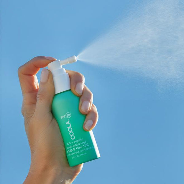 Hand holding and spraying a green bottle of Scalp & Hair Mist Organic Sunscreen SPF 30 | COOLA against a clear blue sky, highlighting skincare and sun protection.