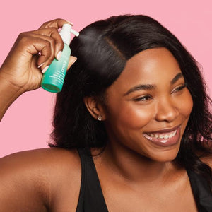 A smiling woman with smooth dark hair applying Scalp & Hair Mist Organic Sunscreen SPF 30 | COOLA near her scalp against a soft pink background.