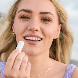 A smiling woman with blonde hair applies Classic Liplux® Organic Lip Balm Sunscreen SPF 30 - Original | COOLA to her lips, promoting skincare and sun protection.