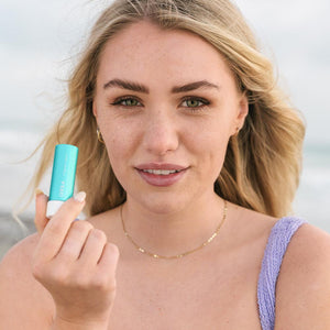 Young woman with natural makeup holding Classic Liplux® Organic Lip Balm Sunscreen SPF 30 - Original | COOLA near her face, promoting skincare and sun protection outdoors.