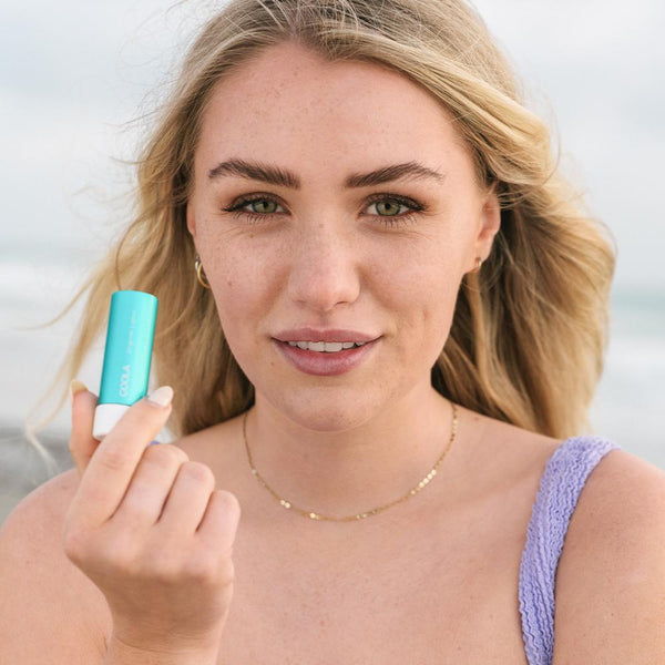 Young woman with natural makeup holding Classic Liplux® Organic Lip Balm Sunscreen SPF 30 - Original | COOLA near her face, promoting skincare and sun protection outdoors.