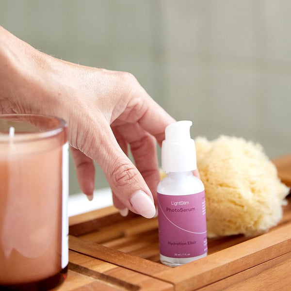 Hand reaching for a bottle of LightStim For Wrinkles | LightStim skincare serum on a wooden tray next to a lit candle and natural sponge, promoting wellness and body care.